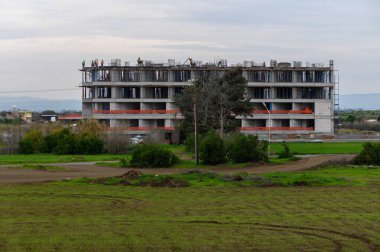 İşçiler tarlalar ve bulutlu bir gökyüzü ile çevrili büyük bir binanın inşasında işbirliği yapıyorlar..