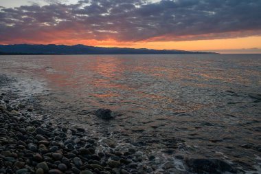 Yumuşak dalgalar taşlara çarpıyor. Canlı renkler gibi. Deniz kenarında sakin bir günbatımında gökyüzünü boyuyor..