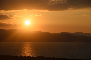 Güneş durgun suların ve yuvarlanan tepelerin üzerinde batarken, gökyüzünü altın renkleri kaplıyor ve huzurlu bir akşam atmosferi yaratıyor..