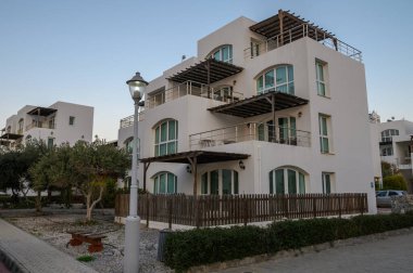 Modern bir tasarıma sahip geniş bir villa, alacakaranlık arka planında yer alan birden fazla balkon ve yeşillik barındırır..