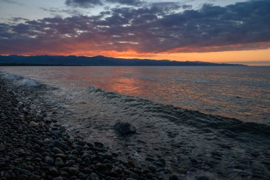 Nefes kesen bir günbatımı, dalgaların sahil boyunca yumuşak çakıl taşlarına hafifçe dokunduğu canlı renklerle gökyüzünü boyar..