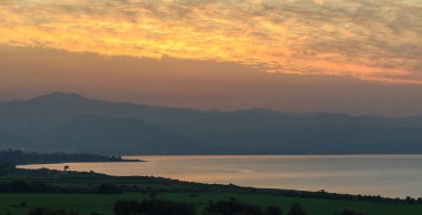 Altın renkler, yumuşak bulutlarla harmanlanır. Güneş, dingin bir göl üzerinde yuvarlanan tepeler arasında batar..