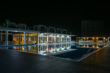 Yumuşak ışıklar sakin suyu yansıtıyor. Konuklar, havuz kenarında sakin bir gecenin atmosferinin tadını çıkarırken..