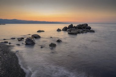 Güneş uzak dağların ardında batarken kayalık plaja doğru yumuşak dalgalar süzülür ve gökyüzünü sıcak renklerle boyarlar..