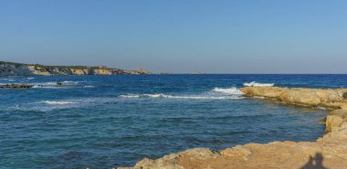 Sakin bir kıyı manzarası, alacakaranlıkta güneşli kayalara karşı parlak mavi suları ve yumuşak dalgaları barındırır..