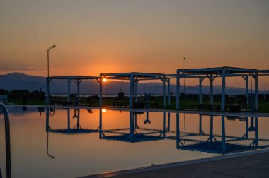 Altın güneş ışığı, uzak dağlar ve alacakaranlıkta sakin yapılar tarafından çerçevelenmiş bir havuzun sakin sularında dans eder..