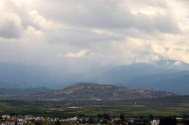 Geniş bir görünüm dalgalı dağları, kırsal bir köyü ve yağmuru tehdit eden karanlık bulutları gözler önüne seriyor..