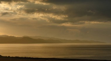 Yumuşak altın renkler, alacakaranlıktaki kasvetli gökyüzüne karşı dağların silüeti gibi sakin suları yansıtır..