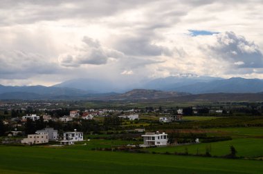 Uzak dağlar ve manzarayı noktalayan modern evler tarafından çerçevelenmiş, dramatik bir gökyüzünün altında geniş yeşil alanlar uzanıyor..