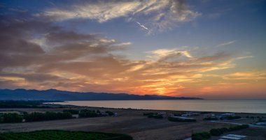 Güneş huzurlu sulara ve yemyeşil tarlalara doğru inerken parlak bir günbatımı göğü sıcak renklerle boyar..