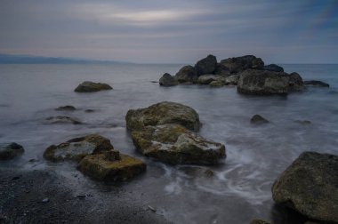 Güneş batarken, pürüzsüz dalgalar kayalık kıyı şeridini sarıp karanlık gökyüzünün altında huzurlu bir ortam yaratıyor..