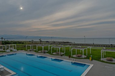 Alacakaranlıkta havuz kenarında, yemyeşil otlar ve okyanus dalgalarının yumuşak sesleriyle çevrili sakin bir ortam..