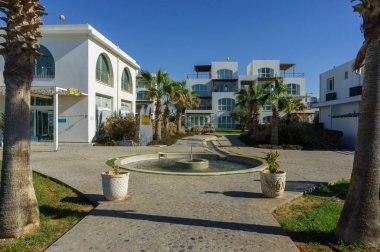 Huzurlu bir tatil köyünde, berrak mavi gökyüzünün altında, huzurlu bir çeşmeyi çevreleyen gür palmiye ağaçları sergilenir..