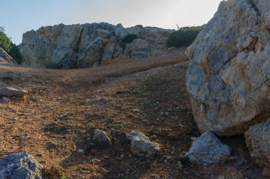 Bir yürüyüşçü, parlak bir güneşin altında, manzaralı bir yamaçta, kayalık zeminde yol alır..