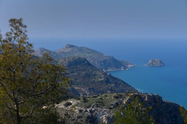 Sakin bir manzara engebeli tepeleri yakalıyor berrak mavi gökyüzünün altında güneş ışığı tarafından öpülen sakin okyanus dalgalarıyla buluşuyor..