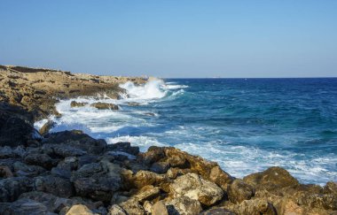 Dinamik dalgalar, berrak mavi gökyüzünün altında engebeli kayalarla çarpışır ve güzel bir kıyı manzarası yaratır..
