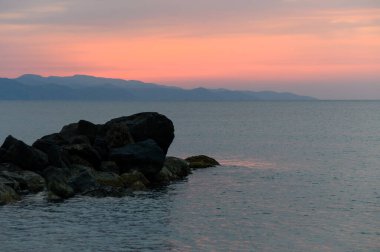 Sakin sular turuncu ve pembe renkleri yansıtır. Dalgalar alacakaranlıkta yavaşça kayalara çarpar.