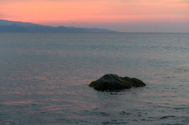 Sakin suların sakin görüntüsü günbatımının yumuşak renklerini yansıtıyor. Issız bir kaya ve uzak dağlarla..