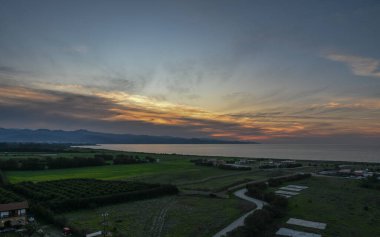 Alacakaranlık, güneş sakin sahil ve yemyeşil tarlalarda batarken gökyüzünü renklendiriyor. Huzurlu bir manzara yaratıyor..