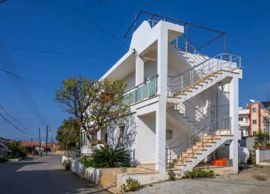 Çağdaş bir bina, açık mavi gökyüzünün altında yeşil ve evlerle çevrili güneşli bir sokakta parlıyor..