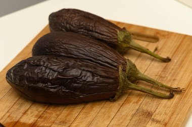 Üç tombul patlıcan, zengin, derin tonlarını ve eşsiz dokusunu sergileyen tahta bir kesme tahtasının üzerinde dinleniyor..