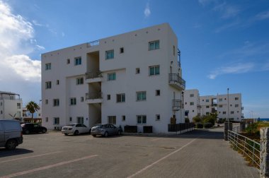 Modern beyaz daireler, yakın park etmiş araçlar ve uzak dalgalarla canlı bir gökyüzüne karşı yükseliyor..