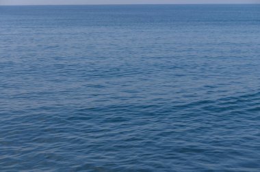 Sakin bir okyanus, sakin mavi gökyüzünü yansıtır. Yumuşak dalgalar suyun yüzeyinde dalgalanmalar oluşturur..