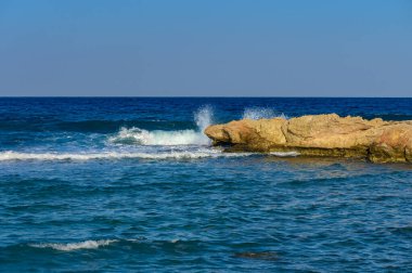 Dalgalar yavaşça kayalık oluşumlara çarpar, güneşli bir günde deniz kenarında sakin bir kıyı atmosferi yaratır..