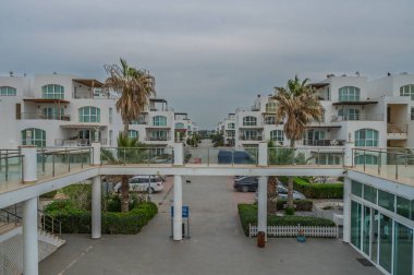 Parlak beyaz binalar palmiye ağaçlarının arasından yükseliyor. Kıyı ufkunda bulutlar sürüklenirken huzurlu bir kaçış yolu sunuyor..