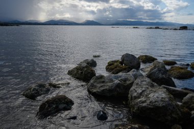 Hafif dalgalar engebeli kayalara çarparken karanlık bulutlar, alacakaranlıkta huzurlu bir su yolu üzerinde beliriyor..