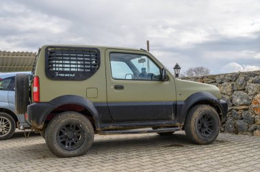 Gaziveren Kıbrıs Rum Kesimi -28.02.2025 zeytin yeşili boyalı kompakt bir off-road aracı bulutlu bir günde desenli taş bir duvarın yanında park edilmiştir..