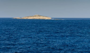 Uzak ada, berrak bir gökyüzü altında barışçıl dalgalarla çerçevelenmiş derin mavi okyanustan doğar..