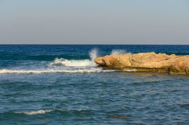 Güneş batarken, yumuşak dalgalar engebeli kayaların üzerinde şakacı bir tur atar ve sakin bir sahil atmosferi oluşturur..