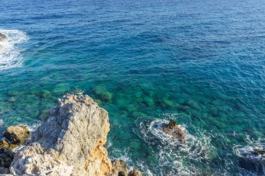 Kristal berrak sular güneşin altında parıldıyor, sakin sahil boyunca engebeli kayaları kucaklıyor..