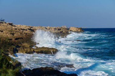 Dalgalar güçlü bir şekilde engebeli kıyı şeridini vurarak sakin mavi denize ve parlak gökyüzüne karşı beyaz köpük oluşturdu..