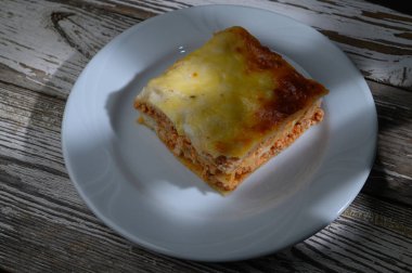 Sağlam bir parça ev yapımı lazanya el değmemiş beyaz bir tabağın üzerinde duruyor, makarna, et ve zengin peynir katmanları sergiliyor. Kırsal ahşap masa rahatlatıcı çekiciliğini vurgular..