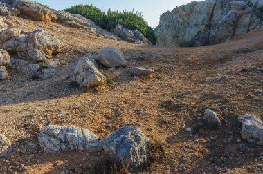 Engebeli bir manzara düz kayalar ve toprak içerir, yemyeşil bitkiler mavi gökyüzünü tamamlar..