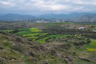 Geniş yeşil alanlar tepe boyunca uzanır, bulutlu gökyüzü ve uzak dağ siluetleri tarafından çerçevelenir..