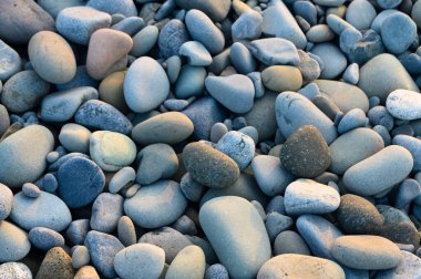 Pürüzsüz ve canlı çakıl taşları sahili kaplıyor. Güneş sakin suların üzerinde batarken renkleri yansıtıyor..