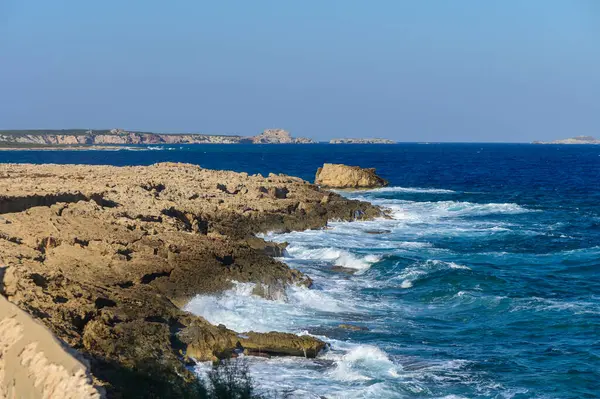 Yumuşak dalgalar dalgalı kıyı şeridine vurur, parlak mavi bir zemin üzerinde güneş ışığını yansıtır..