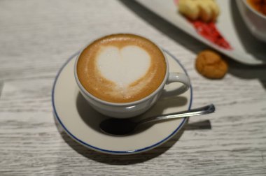 Kalp şeklinde köpük sanatı olan çok güzel işlenmiş bir latte büyüleyici bir kafedeki masanın üzerinde duruyor. Yumuşak ışıklandırma, davetkar atmosferi güçlendirir. Sabah kahvesi ayini için mükemmeldir..