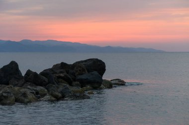 Güneş durgun suların üzerinde batarken, yumuşak pastel renkler gökyüzünü dolduruyor, engebeli kayalar huzurlu manzaraya karışıyor, yansıma ve rahatlama için huzurlu bir atmosfer yaratıyor..