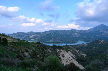 Yuvarlanan tepeler, alacakaranlığın yumuşak tonlarını yansıtan sakin bir gölü beşiyorlar. Kabarık bulutlar havada süzülür, yemyeşil manzaraya gölgeler bırakır. Atmosfer huzurlu, davetkar bir keşif..