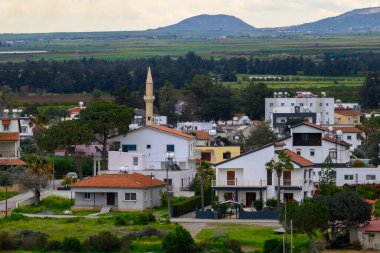 Kıbrıs 'taki huzurlu bir köy, bulutlu bir gökyüzü altında yemyeşil tarlalar ve dağlarla çevrili beyaz yıkanmış evler ve tanınmış bir minareyi gözler önüne seriyor ve yerel cazibe ve sükuneti yakalıyor..