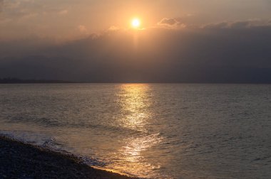 Gün batımında bulutların arasından güneş ışığı doğar, sakin denize altın bir parıltı saçar. Çakıl taşlarıyla kaplı kıyı şeridi, arka planda gün geceye dönerken huzurlu bir kaçış sunuyor..