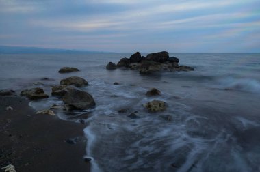 Alacakaranlık çöktüğünde dalgalar engebeli bir kıyı şeridine hafifçe vurarak okyanusun dingin sularına kısmen batmış kayaları ortaya çıkararak alacakaranlık renklerini harmanlıyor..
