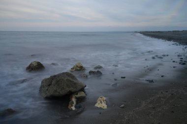 Alacakaranlık çöktüğünde yumuşak dalgalar kasvetli bir gökyüzünün altında kayalık sahili okşar. Kıyı şeridi genişliyor, toprak ve suyun bir karışımını sergiliyor, huzurlu bir tefekküre yol açıyor..