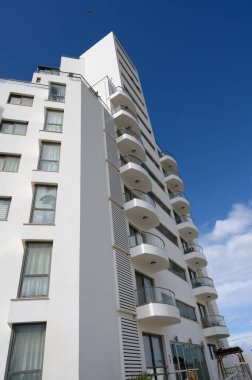 Uzun, çağdaş bina parlak bir gökyüzüne karşı görkemli bir şekilde duruyor. Balkonlar zarafet ekler, canlı bir şehir ortamındaki mimari güzelliği vurgular..