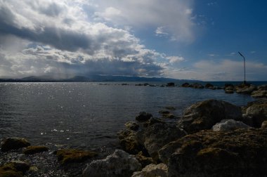 Alacakaranlıkta kıyı şeridi boyunca kayalara yumuşak dalgalar çarpar. Uzak tepelerin üzerinde kara bulutlar belirirken güneş suyun yüzeyine canlı yansımalar saçar..