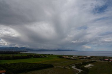 Huzurlu bir kıyı manzarası, sakin denizle buluşan yumuşak tepeler ve tarlalarla gözler önüne seriliyor. Yukarıdaki dönen bulutlar yaklaşan bir değişimi işaret ederken, uzak dağlar manzaraya derinlik katıyor..